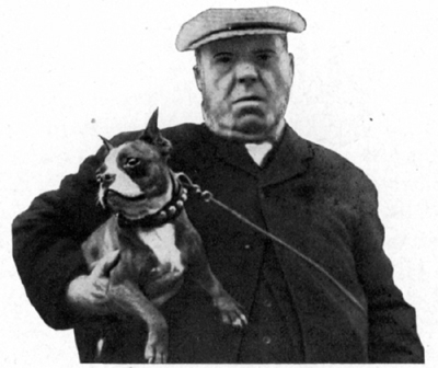 A gentleman in a cap holds a dog under one arm.