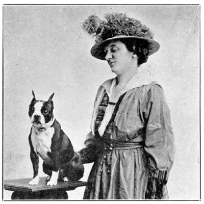 A woman with a rather large hat stands next to a female sitting on a table