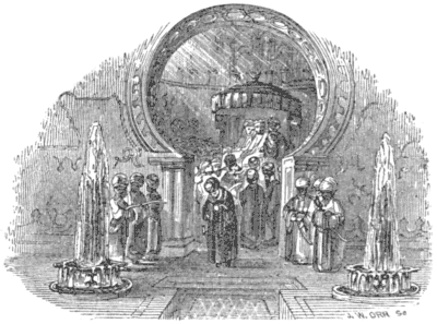 A man walks, head bowed, through an arch, with three men flanked on either side of it,
while other men are in a group behind him