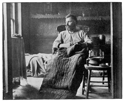 A photograph of a man sitting in a chair, with his knees covered with a blanket.