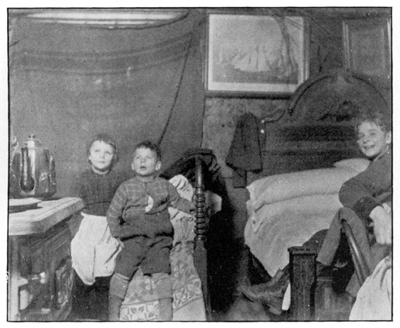 A crowded room with three children, two beds and a stove.