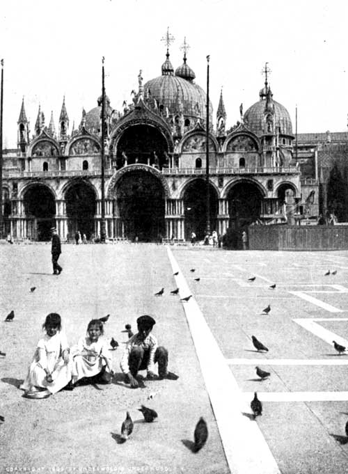 Children feeding Pigeons in the Piazza of St. Mark, Venice. Notice the three flag-poles, and the bronze horses over the central doorway of the Cathedral.  