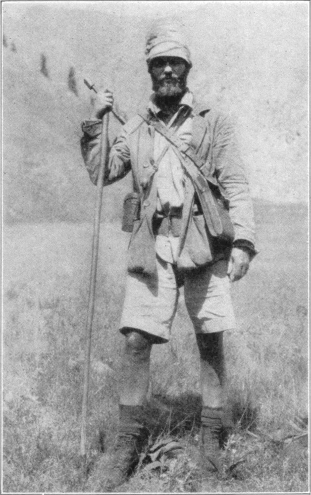 Kermit Roosevelt in hunting-kit with a long walking stick, with a binocular case around his neck and a rifle on his back.
