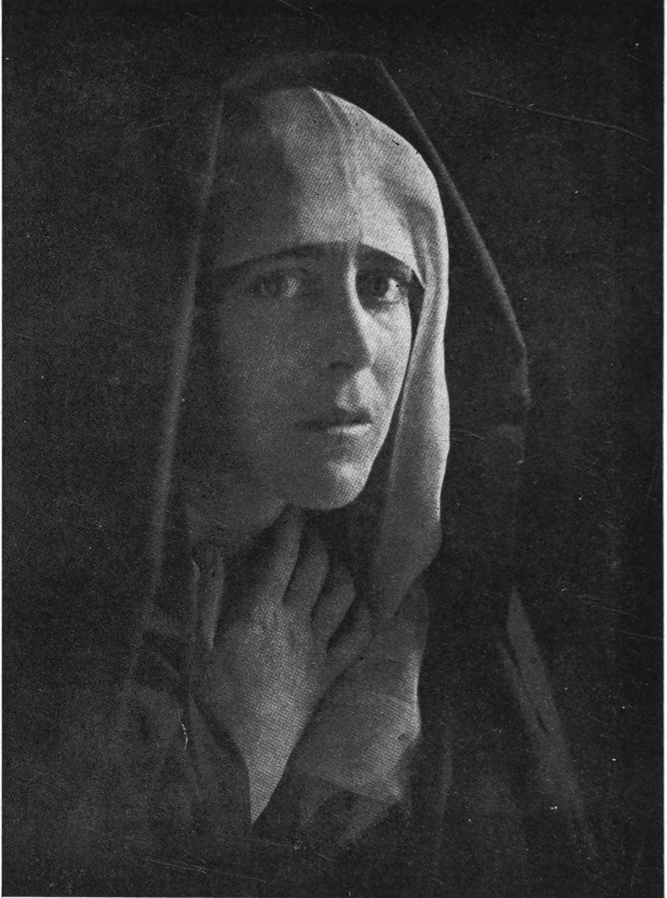 Photo of the face of a woman, lit from the side; she wear a cloth head covering and holds her right hand at her throat. Darkness enshrouds the image.