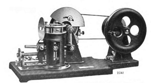 A rotating cylinder mechanism on a wooden base, connected by belt to a large four-spoked flywheel.