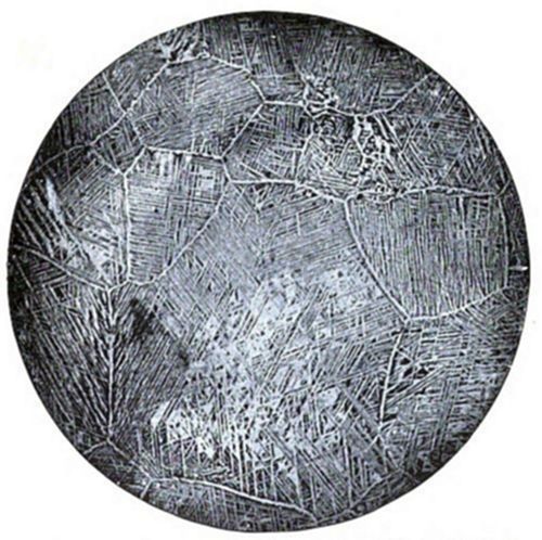 Circular micrograph showing a Widmanstätten-like microstructure — needle-like or plate-like crystalline patterns arranged within irregular grain boundaries, typical of certain metal alloys viewed under a microscope.