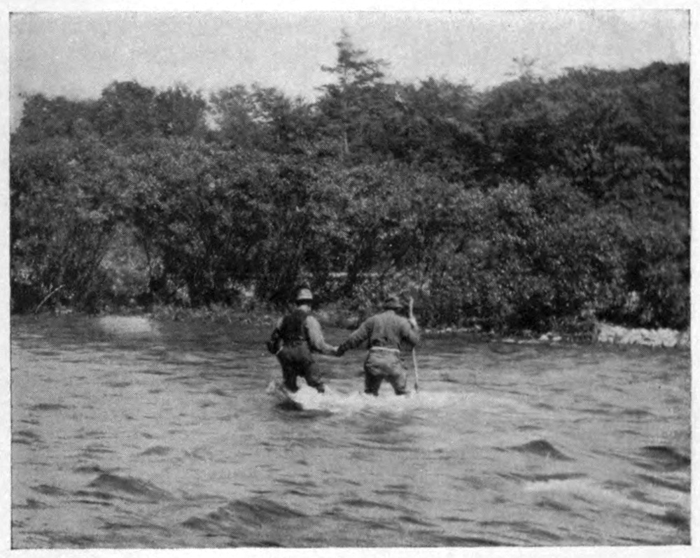 Photo of two men wading a rapid river with water to their thighs.