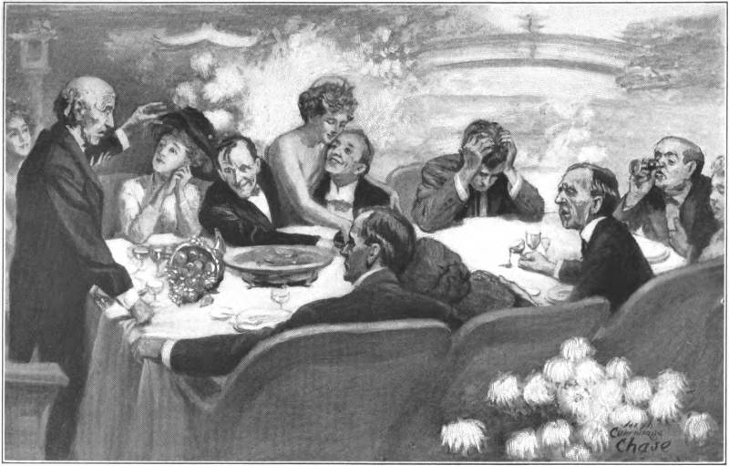 A group of men and women around a dinner table. Most are looking towards a man standing at one end who appears to be speaking.