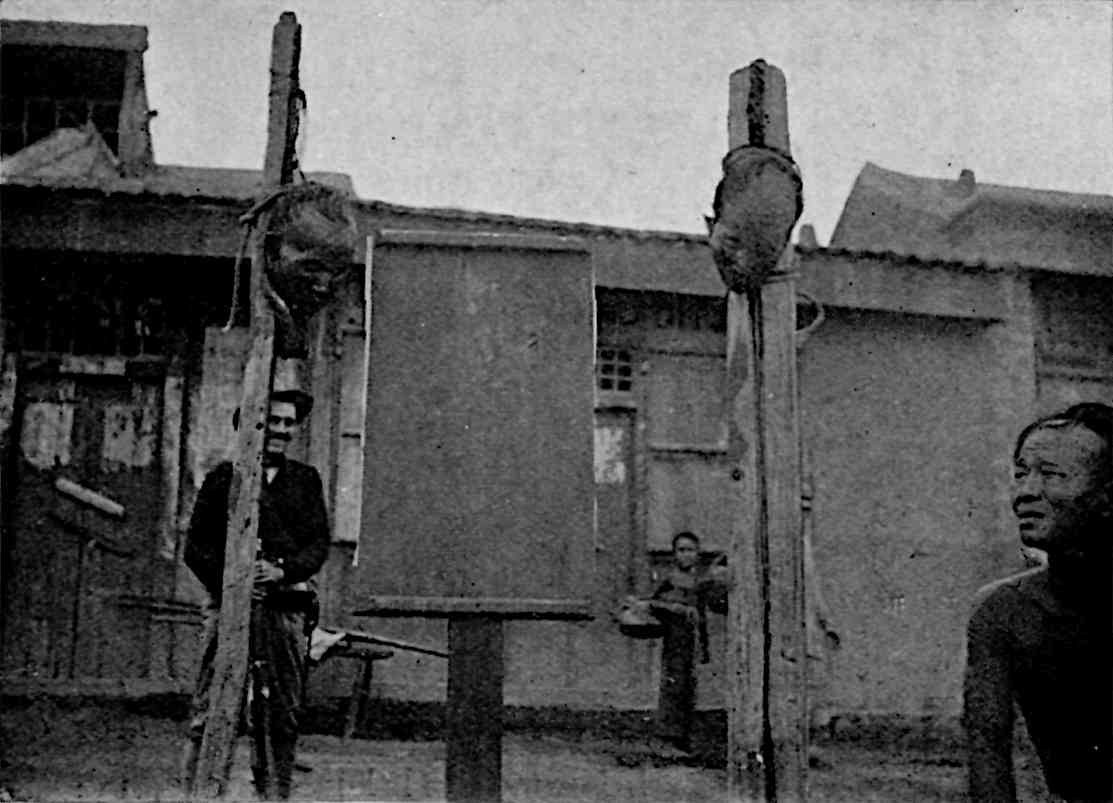 Man on right looking at 2 decapitated heads hanging from poles in the center