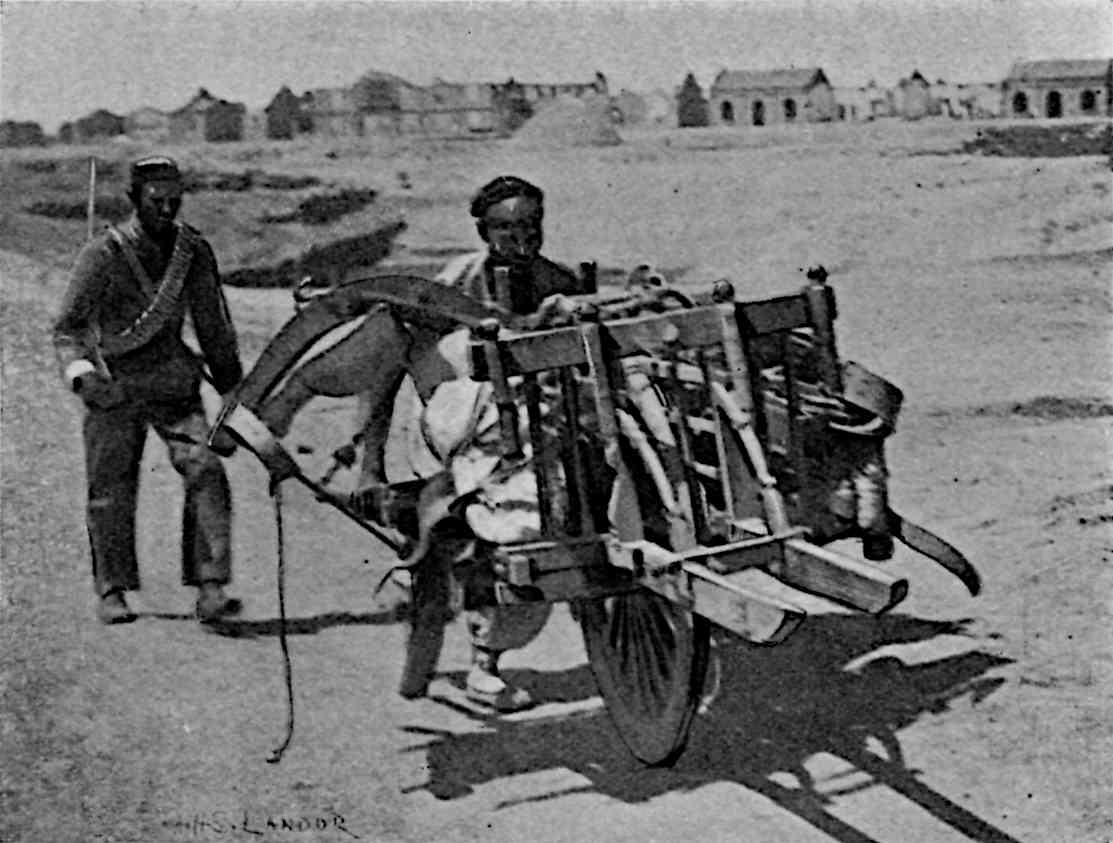 1 person pushing single-wheeled wheelbarrow with another behind
