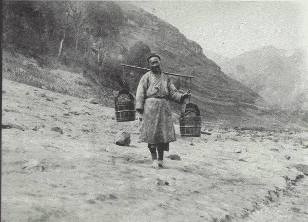 THE UBIQUITOUS WATER CARRIER

Drawing the water and hewing the wood are daily chores in China, mostly
carried out by women—though this is a picture of a man, a half-wit