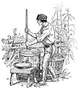 A man operates a barrel-sized mill by holding a long stick. The corn is coming out of the side of the barrel.