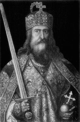 A portrait of Charlemagne. He has long hair and a beard, and wears richly
embroidered clothes and a jewelled and decorated crown. He holds an orb with a cross
in his left hand, and a raised sword in his right.