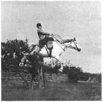 Man on a grey horse jumping a fence in a side-saddle.