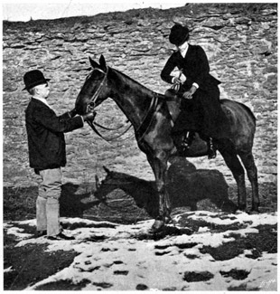 A mounted woman preparing to dismount while a groom hold the horse's head