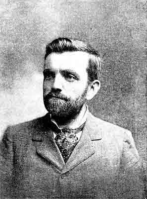 Trimmed beard and moustache; square standup collar, tied patterned ascot, small check jacket.