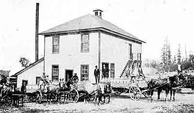 Two story frame building; four teams of horses hitched to wagons full of metal milk cans.