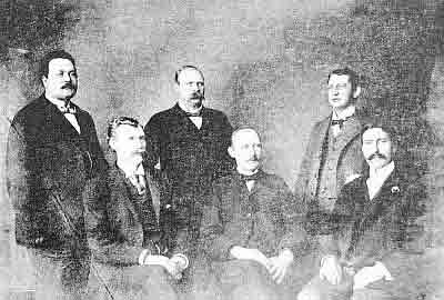 Six men, three sitting, three standing behind, in variety of previously described men's dress.