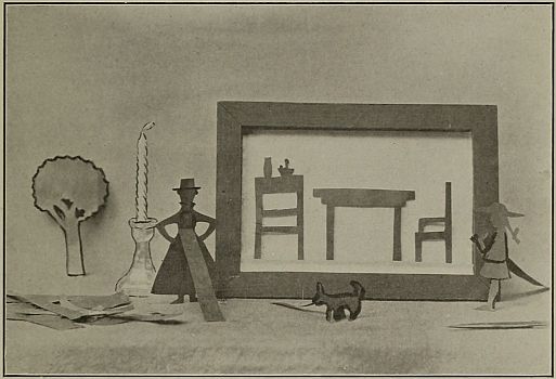 photograph of sillouette scen of table, chair, sideboard, additional pieces outside of frame, two dolls, a dog a tree, a candle