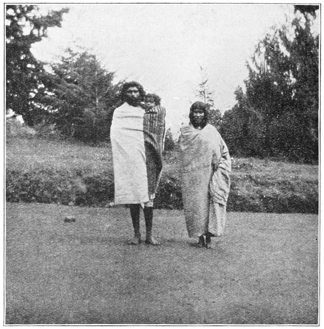 FIG. 9.—A TODA MAN, SIRIAR (20), WITH HIS WIFE AND CHILD, SHOWING THE ORDINARY METHOD OF WEARING THE ‘PUTKULI.’