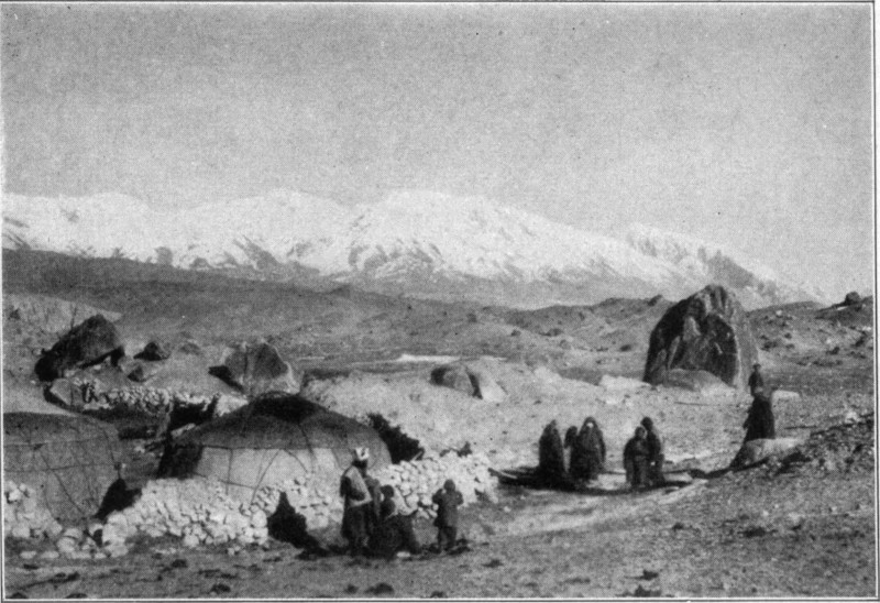 Two yurts in a barren landscape with people in the foreground and mountains in the background.