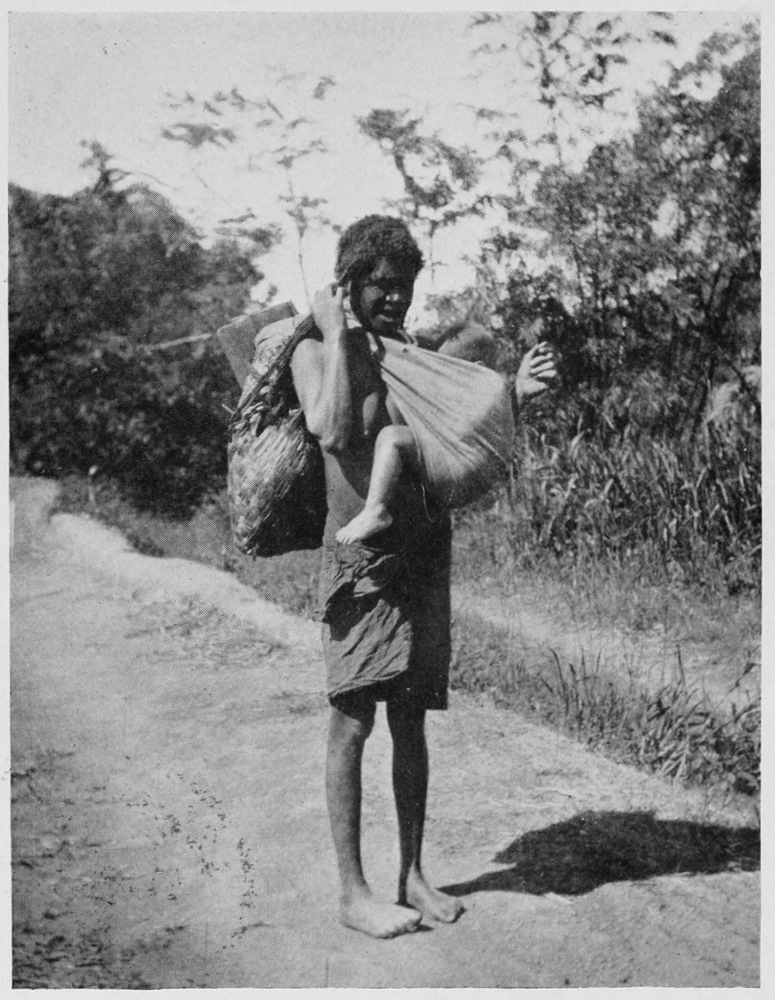 Barefoot young woman on a dirt road. She carries a young child in a sling across her chest, and holds a bag of supllies over her right shoulder